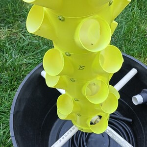 May include: A yellow plastic vertical garden tower with multiple planting holes. The tower is sitting in a black plastic container with a white pipe and black tubing. The pipe has the text "73&deg;F [ASTM D-1785 [N] OK IA SL-M 05/08/0" written on it. The tower has the letter "B" written on it.