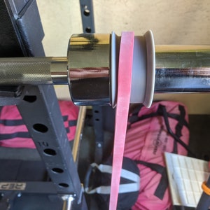May include: A weightlifting barbell with a pink resistance band attached. The band is looped around a silver collar and extends downwards. The barbell is mounted on a black weight rack, with gym bags in the background.