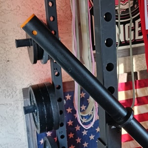 以下が含まれることがあります： 黒いウェイトリフティングラックにバーベルとウェイトプレートが置かれています。ラックには黒とオレンジのプルアップバーが取り付けられています。ラックはアメリカ国旗の前にあります。