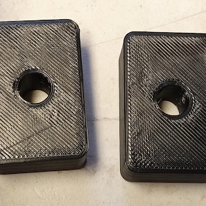 May include: Two black rectangular blocks with a circular hole in the center. The blocks have a textured surface with diagonal lines. The objects appear to be 3D printed.