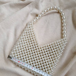 May include: A white beaded handbag with a top handle. The bag is made of small, round pearls and is approximately 20 inches in length.