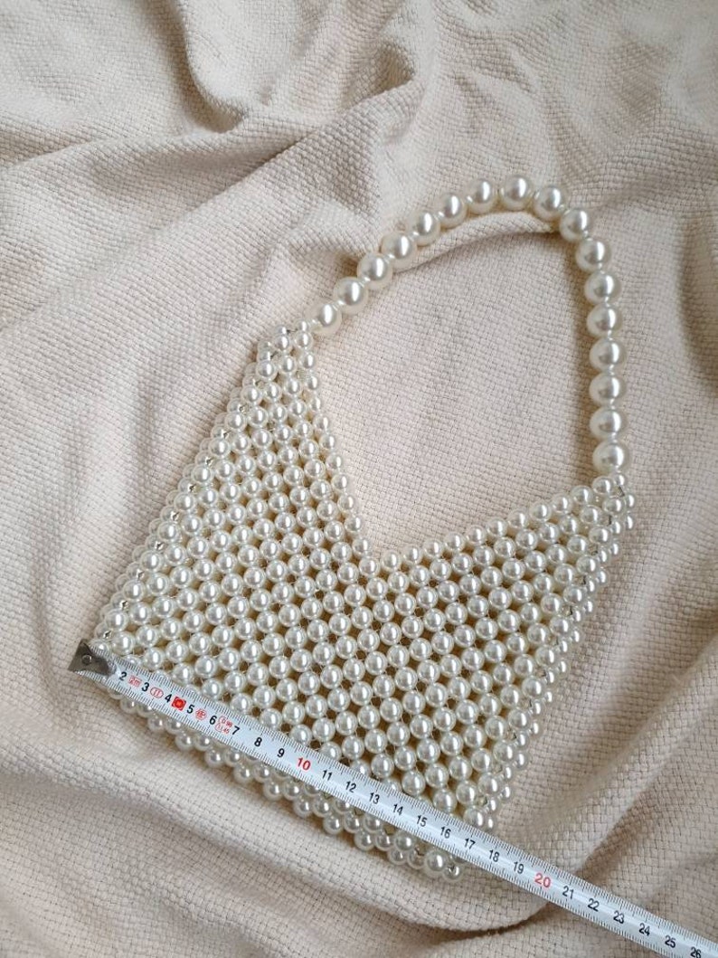 May include: A white beaded handbag with a top handle. The bag is made of small, round pearls and is approximately 20 inches in length.