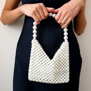 May include: A white pearl beaded handbag with a triangular shape. The bag has a top handle made of pearls.