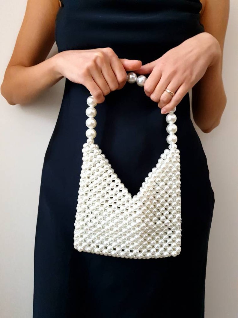 May include: A white pearl beaded handbag with a triangular shape. The bag has a top handle made of pearls.