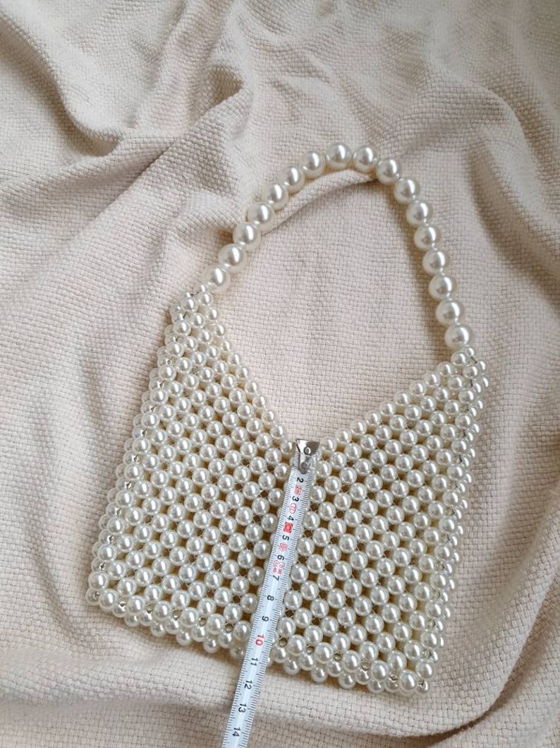 May include: A white pearl beaded handbag with a top handle. The bag is approximately 11 inches wide.