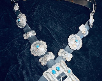 May include: A black leather belt featuring a large rectangular silver buckle adorned with four turquoise stones. The belt strap is decorated with multiple circular and scalloped silver concho plates, each centered with a small turquoise stone.