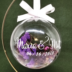 May include: A clear glass ornament with dried purple flowers inside, suspended by a white ribbon. The ornament has the names "Maria & Matt" and the date "06/16/2018" in white script. The ornament hangs from a gold metal stand.