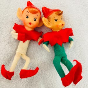 Puede incluir: Dos figuras de duendes vintage. Uno tiene un cuerpo color crema, sombrero y zapatos rojos, y un cuello rojo. El otro tiene un cuerpo verde, sombrero y zapatos rojos, y un cuello rojo. Ambos tienen caras pintadas con orejas grandes.