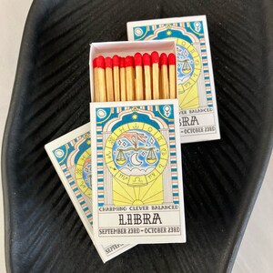 May include: A box of matches with a blue and yellow design featuring the astrological sign Libra. The text on the box reads "Charming Clever Balanced Libra September 23rd - October 23rd".