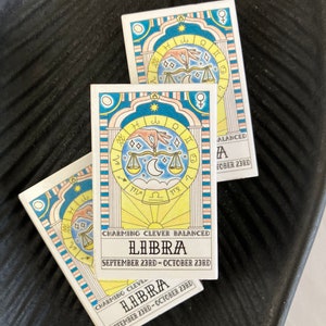 May include: Two illustrated cards with a blue, yellow, and pink color scheme. The cards feature a Libra zodiac symbol with the text "CHARMING CLEVER BALANCED LIBRA SEPTEMBER 23RD - OCTOBER 23RD".