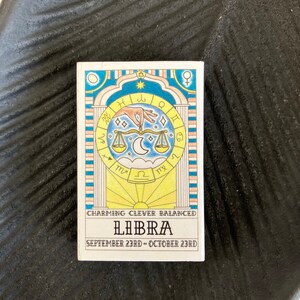 May include: A colorful illustration of the Libra zodiac sign with the dates September 23rd - October 23rd. The illustration features a scale, a hand, and astrological symbols. The text "Charming, Clever, Balanced" is written below the illustration.