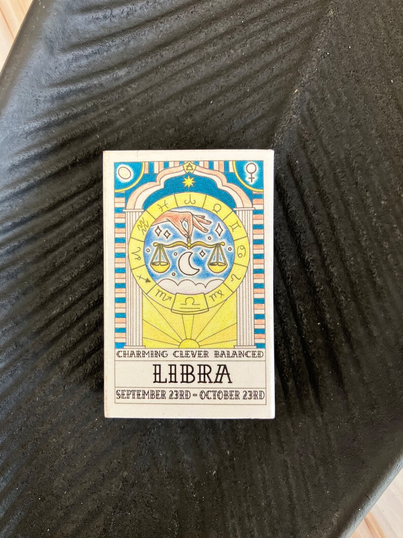 May include: A colorful illustration of the Libra zodiac sign with the dates September 23rd - October 23rd. The illustration features a scale, a hand, and astrological symbols. The text "Charming, Clever, Balanced" is written below the illustration.