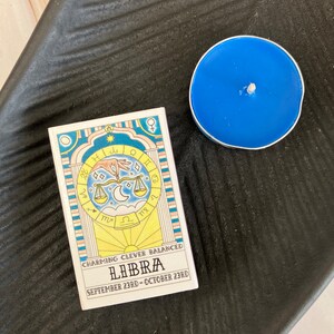 May include: A white card with a yellow, blue, and pink illustration of a Libra zodiac sign. The card reads "Charming, Clever, Balanced, Libra, September 23rd - October 23rd".