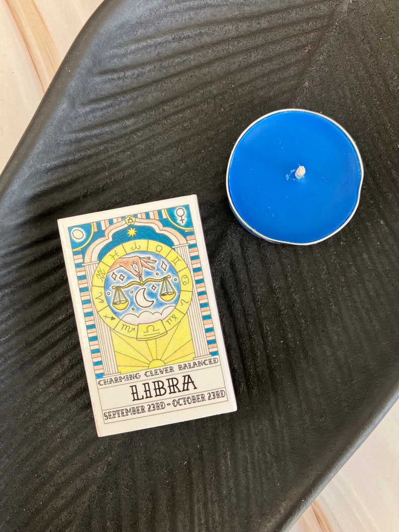May include: A white card with a yellow, blue, and pink illustration of a Libra zodiac sign. The card reads "Charming, Clever, Balanced, Libra, September 23rd - October 23rd".