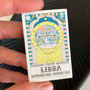 May include: A white card with a colorful illustration of a Libra zodiac sign. The card has the text "CHARMING CLEVER BALANCED LIBRA SEPTEMBER 23RD - OCTOBER 23RD".