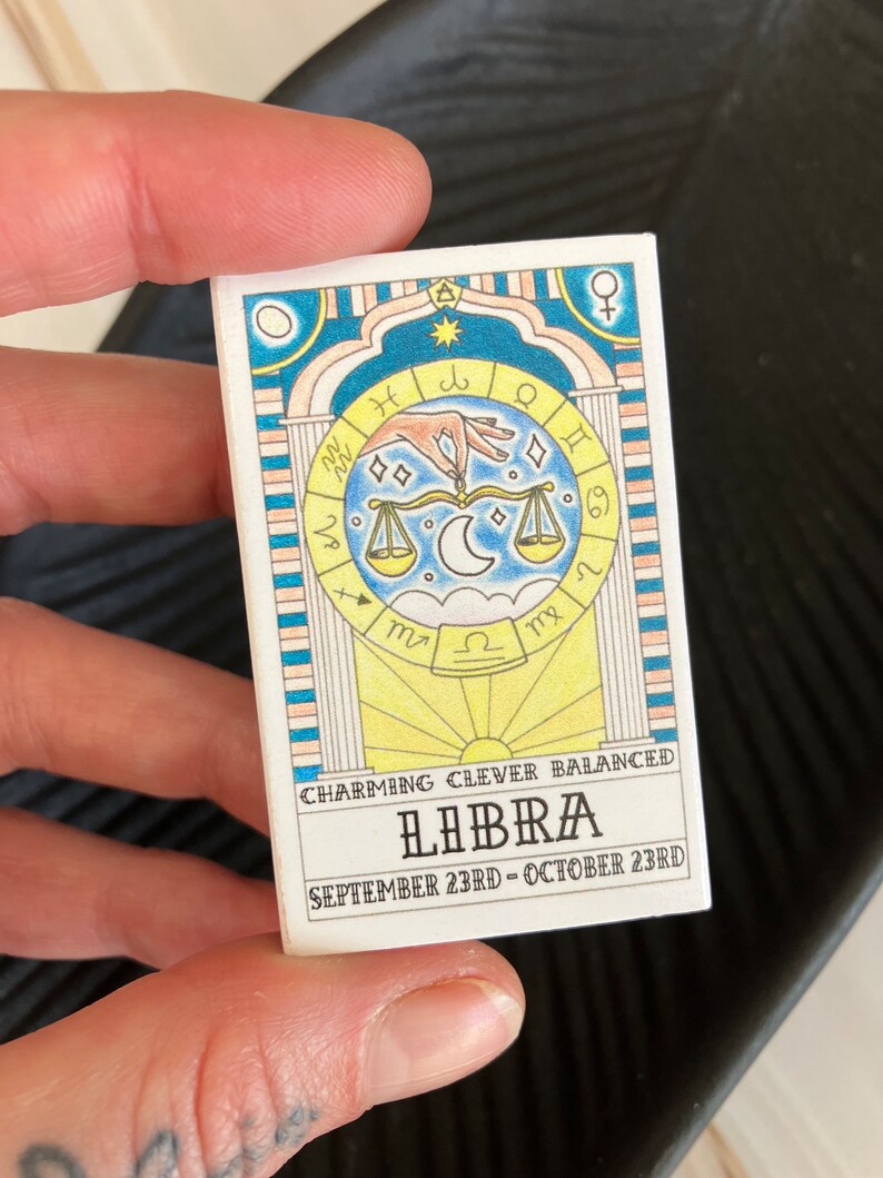 May include: A white card with a colorful illustration of a Libra zodiac sign. The card has the text "CHARMING CLEVER BALANCED LIBRA SEPTEMBER 23RD - OCTOBER 23RD".