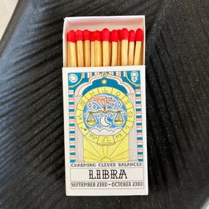 May include: A box of matches with a Libra zodiac sign design. The box is white with blue and yellow accents. The matches have red tips. The text on the box reads "Charming Clever Balanced Libra September 23rd - October 23rd".
