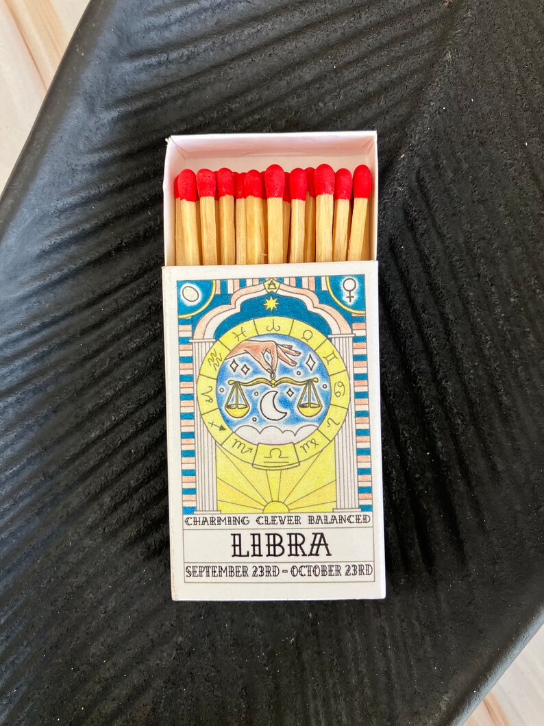 May include: A box of matches with a Libra zodiac sign design. The box is white with blue and yellow accents. The matches have red tips. The text on the box reads "Charming Clever Balanced Libra September 23rd - October 23rd".