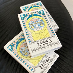 May include: Two white matchboxes with a yellow, blue, and pink striped border. The matchboxes feature a zodiac sign illustration of Libra with the text "CHARMING CLEVER BALANCED LIBIRA SEPTEMBER 23RD - OCTOBER 23RD".