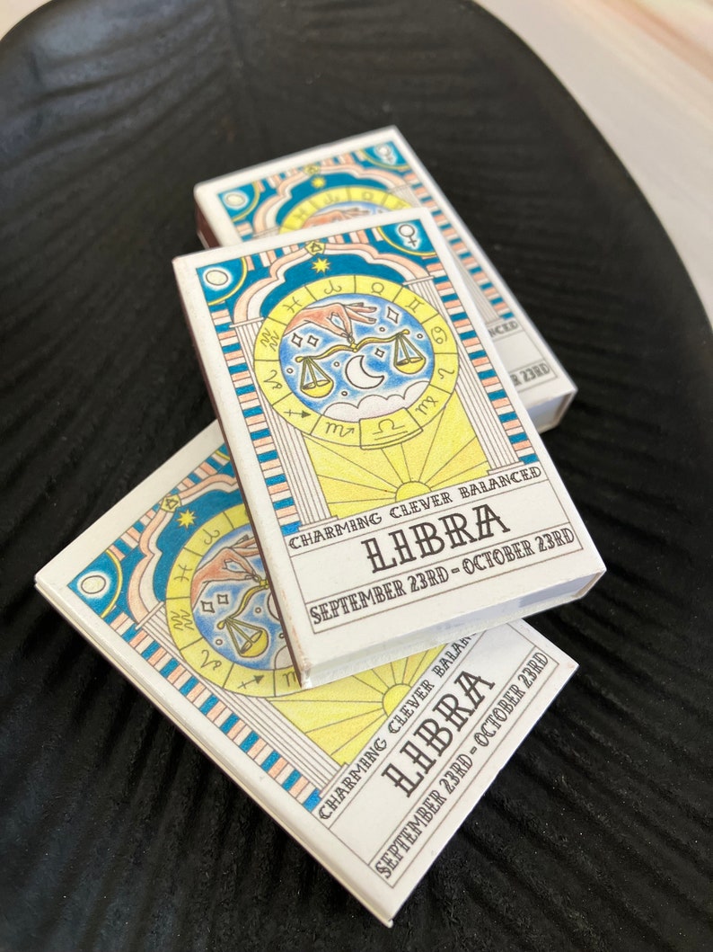 May include: Two white matchboxes with a yellow, blue, and pink striped border. The matchboxes feature a zodiac sign illustration of Libra with the text "CHARMING CLEVER BALANCED LIBIRA SEPTEMBER 23RD - OCTOBER 23RD".