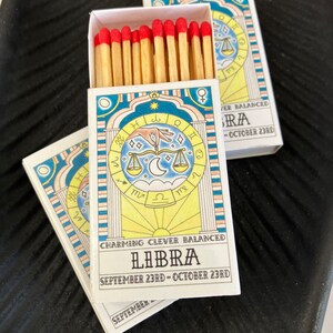 May include: A box of matches with a blue, yellow, and white design featuring the astrological sign Libra. The box has the text "Charming Clever Balanced Libra September 23rd - October 23rd".
