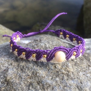 Könnte beinhalten: Lila-weißes Makramee-Armband mit goldfarbenem Perlenakzent und einer einzelnen rosa Muschelperle.