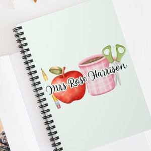 Può includere: Un quaderno a spirale verde con una copertina verde chiaro che presenta una mela rossa, una tazza a scacchi rosa e bianca, una matita gialla e delle forbici verdi. Il testo "Mrs. Rose Harrison" è scritto in corsivo nero.