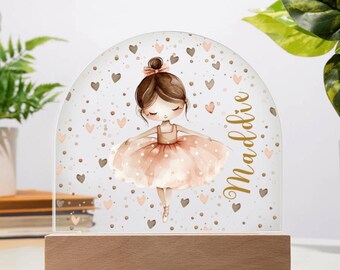Może przedstawiać: Dekoracyjna lampka nocna z akwarelową ilustracją baletnicy w różowej tutu. Łukowaty panel akrylowy jest ozdobiony sercami i kropkami, a imię "Maddie" jest napisane złotym pismem. Podstawa wykonana jest z jasnego drewna.