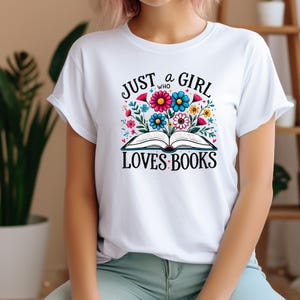 Può includere: Maglietta bianca con un design grafico di un libro aperto con fiori e il testo "Just a girl who loves books".