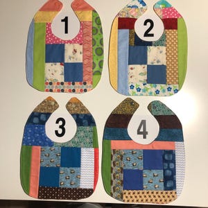 May include: Four handmade patchwork baby bibs. Each bib features a unique combination of patterned fabrics in various colors, including blue, green, pink, and brown. The bibs are numbered 1 through 4, with a curved shape and a snap closure at the neck.