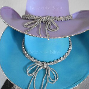 May include: Two cowboy hats stacked on top of each other. The top hat is lavender with a silver rhinestone trim and a silver rhinestone bow. The bottom hat is turquoise with a silver rhinestone trim and a silver rhinestone bow. The text "Belle of the Bling" is printed on both hats.