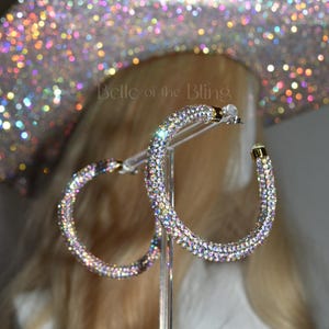 May include: A pair of large hoop earrings with a gold tone setting. The hoops are completely covered in iridescent rhinestones.