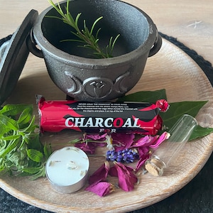 May include: A black cast iron cauldron with a triple moon design, a pack of charcoal briquettes, a small glass vial, a tea light candle, and dried herbs and flowers on a wooden tray.