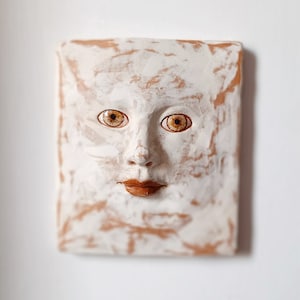 Puede incluir: Una escultura de pared de cerámica blanca con una cara, ojos marrones y una boca marrón. La escultura tiene una superficie texturizada y está pintada con pintura blanca y marrón.