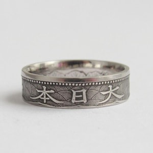 May include: A silver ring with Japanese characters engraved around the band. The ring has a detailed pattern of waves and dots. The ring is set against a white background.