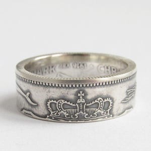 Könnte beinhalten: Ein silberner Ring mit einem detaillierten Kronenmuster. Der Ring scheint aus einer Münze gefertigt zu sein, mit sichtbarem Text und komplizierten Mustern um das Band. Der Ring ist vor einem schlichten weißen Hintergrund platziert.