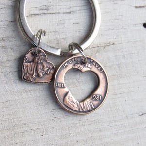 May include: A copper-colored keychain with two charms. One charm is a heart-shaped pendant with a detailed design. The other is a coin with a heart-shaped cutout and the year 1981. The keychain is on a silver metal ring.