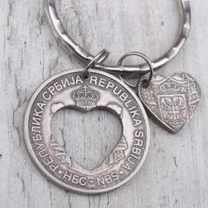 Puede incluir: Un llavero plateado con una moneda con un recorte en forma de corazón y texto en cirílico. Un pequeño dije en forma de corazón con un emblema cuelga. El llavero está unido a un anillo plateado.