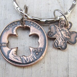 May include: A copper penny keychain with a four-leaf clover cut out. The penny is dated 1981 and has the words "In God We Trust" and "Liberty" visible. The keychain also features a smaller four-leaf clover charm.