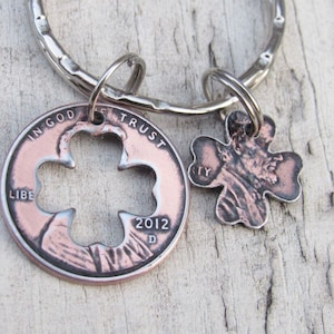Puede incluir: Un llavero de plata con un dije de centavo de cobre con un trébol de cuatro hojas recortado y el texto "In God We Trust" y "Liberty" y un dije de trébol de cuatro hojas de cobre con el año "2012" en el dije de centavo.