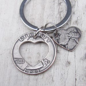 May include: Silver key chain with a heart-shaped cutout in a 1981 quarter. The quarter has the words "LIBERTY" and "IN GOD WE TRUST" inscribed on it. A small heart-shaped charm with a woman's profile is attached to the key chain.