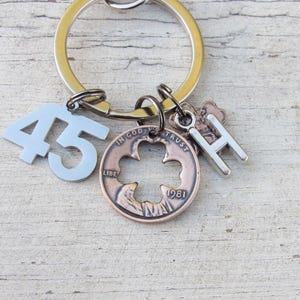 May include: A silver keychain with multiple charms. Charms include a light blue number 45, a copper coin with a cross-shaped cutout, a silver letter "H", and a copper coin with the text "IN GOD WE TRUST" and the year "1981".