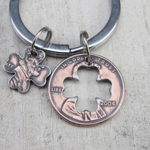 Könnte beinhalten: Ein silberner Schlüsselanhänger mit zwei vierblättrigen Kleeblatt-Anhängern aus Kupfer. Ein Anhänger ist ein US-Penny mit den sichtbaren Worten "In God We Trust" und "Liberty". Der andere Anhänger ist ein kleineres vierblättriges Kleeblatt mit dem sichtbaren Wort "RT". Der Penny-Anhänger hat ein vierblättriges Kleeblatt, das aus der Mitte herausgeschnitten ist. Das Jahr 2004 ist auf dem Penny-Anhänger sichtbar.
