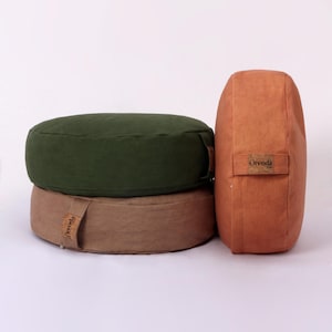 May include: Three meditation cushions in shades of green and brown. The round cushions are stacked, with a taller, rectangular cushion to the right. Each cushion has a small tag that reads "Orenda".