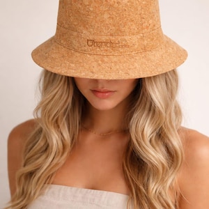 Peut inclure: Un chapeau de seau en liège marron clair avec un large bord. Le chapeau porte le mot "Orenda" imprimé sur le devant. Le chapeau est fait d'un matériau texturé. Une personne aux cheveux blonds porte le chapeau.