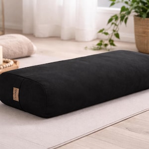 May include: A black rectangular yoga bolster with rounded ends. The bolster is made of a soft, textured fabric and has a small, rectangular tag on one end. It rests on a light gray yoga mat.