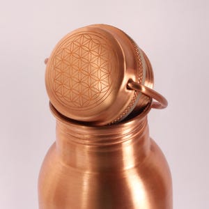 May include: A copper water bottle with a lid featuring an engraved flower of life design. The bottle has a polished finish and a rounded shape.