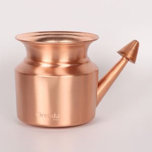 May include: A copper-colored neti pot with a rounded body, flared rim, and a spout. The spout is angled and has a conical tip. The pot has the text "Orenda India" printed on the side. The pot is designed for nasal irrigation.