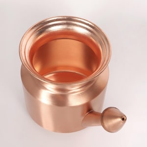 May include: A copper-colored neti pot, a nasal irrigation device, is shown from a top-down perspective. The pot has a rounded body, a wide opening, and a spout with a small tip. The copper surface reflects light, highlighting its shape.