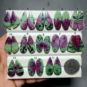 Op de afbeelding: Een collectie van dertien paren gepolijste robijn in zoisiet cabochons gerangschikt op een wit displayblad, genummerd van 1 tot 13. Elke steen toont een mix van levendig magenta en gevlekt groen met zwarte insluitingen, naast een muntstuk.